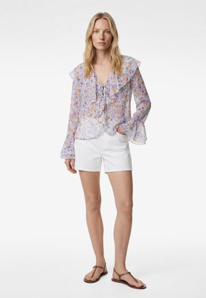 Kobieta w przezroczystej, kwiecistej bluzce z falbanami i długimi rękawami, białych szortach oraz brązowych, płaskich sandałach z ćwiekami, stojąca z jedną ręką w kieszeni.