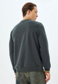 Sudadera de pullover en tela gris oscuro con puños y dobladillo acanalados. Presenta un acento de rayas amarillas y blancas en el cuello. Pantalones de mezclilla azules visibles.
