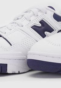 Białe buty sportowe z cholewką ze skóry, fioletowymi akcentami, perforowanymi detalami i teksturowaną gumową podeszwą. Sznurowadła są białe.