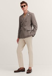 Grau gestreifter Doppelreiher-Blazer, beigefarbene Slim-Fit-Hosen und braune Mokassins. Accessories umfassen dunkle Sonnenbrillen und eine weiße Reversblume.