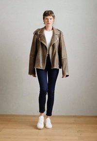 Καφέ δερμάτινο oversized μπουφάν με ασημένια αξεσουάρ, φορεμένο πάνω από ένα λευκό τοπ και σκούρα στενά τζιν, σε συνδυασμό με λευκά αθλητικά παπούτσια σε ανοιχτό ξύλινο δάπεδο.