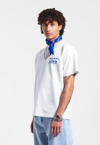 Bílý bavlněný tričko s modrým grafickým textem. Model má kolem krku modrou šátek a světlé modré džíny. Stříbrné šperky doplňují outfit.