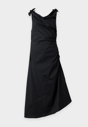 Femme portant une robe midi noire sans manches avec un ourlet asymétrique et des bretelles nouées aux épaules, portée avec des sandales noires ouvertes à talons bas.