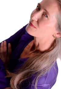 Porträtt av en person med långt, rakt grått hår, iförd en lila skjorta med knappar, som visar en ljus hudton och guldörhängen.