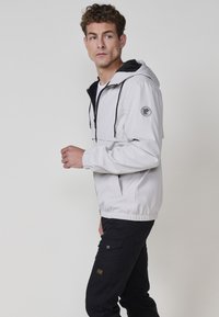 Giacca impermeabile grigio chiaro con accenti neri, cappuccio, polsini elasticizzati e tasca frontale. Abbinata a pantaloni cargo neri con tasca applicata.
