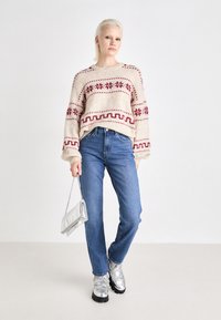 Πλεκτό πουλόβερ σε κρέμα με μαρουνί γεωμετρικά σχέδια, συνδυασμένο με μπλε τζιν και ασημένια παπούτσια με πλατφόρμα, κρατώντας μια ασημένια τσάντα.