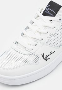 Hvide sneakers med perforeret læder, flade hvide snørebånd og en sort mærketunge. Gummisål med stikningsdetaljer. Signatur på siden.