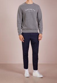 Hombre vestido con sudadera gris de Hackett, pantalones azul marino, calcetines blancos y zapatillas blancas, de pie contra un fondo rosa pálido.