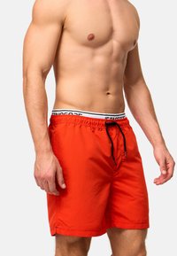 Oberkörperfreier Mann trägt leuchtend rote Badeshorts mit schwarzer Kordel und sichtbarem weißen Bund mit der Aufschrift "JEANSCORP" vor einfarbigem Hintergrund.