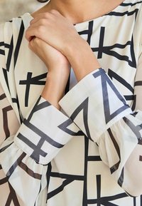 Sheer long-sleeve blouse featuring a geometric black pattern on a white background, with gathered cuffs and a round neckline.