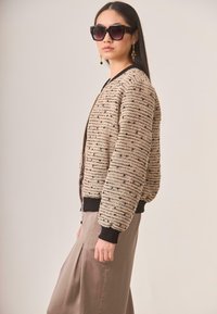 Chaqueta bomber texturizada en beige con detalles en negro y patrones a rayas. Luciendo una falda marrón claro, la modelo se encuentra de lado, complementada con gafas de sol oscuras.