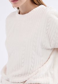 Suéter blanco de textura, con diseño de trenzas y cuello en forma de ondas. Material suave y peludo con un corte holgado.