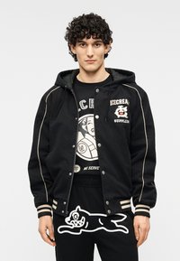 Μαύρο μπουφάν στυλ φούτερ με λευκές λεπτομέρειες. Διαθέτει κουμπιά και πλαϊνές τσέπες, και φοριέται πάνω από μαύρη και λευκή T-shirt με γραφικό σχέδιο.