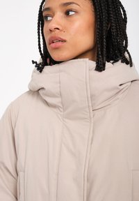 Beige puffer-jakke med en høj krave, der har synlige sømme og en justerbar hætte. Den har to sidelommer.