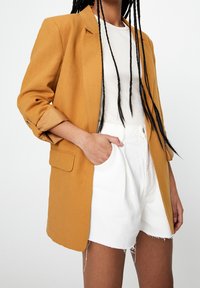 Blazer oversize jaune moutarde avec manches retroussées, détail de bouton unique, complété par un haut blanc ajusté et un short en denim blanc.
