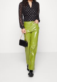 Blouse noire transparente avec un motif de pois dorés, associée à un pantalon taille haute vert citron brillant. Sac à main noir matelassé et bottes épaisses.