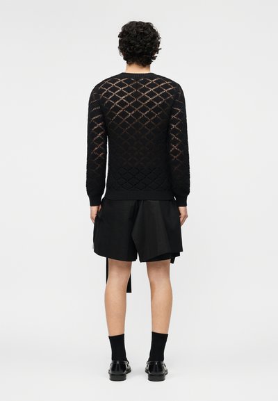 Άνδρας που στέκεται, φοράει μαύρο ανοιχτό πλεκτό πουλόβερ με μακριά μανίκια, φαρδιά μαύρα σορτς, μαύρες κάλτσες και μαύρα παπούτσια, γυρισμένος προς την αντίθετη κατεύθυνση από την κάμερα.