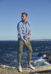 Lichtblauw shirt met een bloemenpatroon, olijfgroene cargobroek en witte sneakers. Staande op een boomstam nabij de oceaan, heldere blauwe lucht.