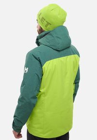 Veste verte et turquoise avec capuche, présentant un motif à carreaux et des blocs de couleur unie. Le bonnet assorti est d'un vert vif avec un texte logo.
