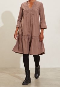 Femme portant une robe mauve jusqu'aux genoux avec de la broderie, des leggings noirs et des bottines à lacets noires marchant sur un sol clair avec un fond de mur uni.