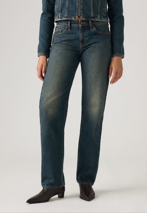 Person iført straight-leg blå jeans, matchende blå denimjakke og sorte spidse ankelstøvler står på en hvid baggrund.