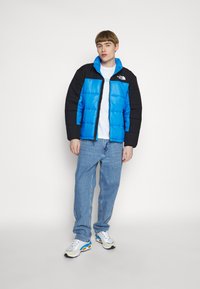 Blå og sort pufferjakke med lynlås, der har høj krave og logo, båret over en hvid t-shirt og lyseblå jeans.