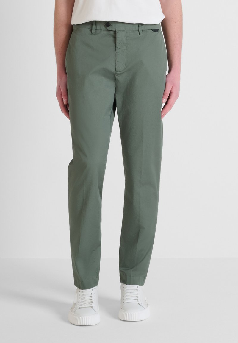 Pantaloni in cotone verde con vestibilità su misura, dotati di chiusura con zip e tasche frontali, abbinati a sneakers bianche. Tessuto liscio, resistente alle pieghe.