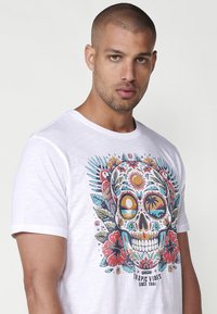 Balta trumpa rankovė t-shirt su spalvinga kaukolės dizainu, kuris apima gėlių elementus, palmės ir paukščius, atspausdinta ant minkštos medžiagos.