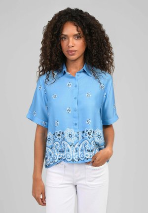 Femme aux cheveux bouclés portant une chemise bleue à manches courtes avec des motifs floraux et un pantalon blanc, debout devant un fond uni.