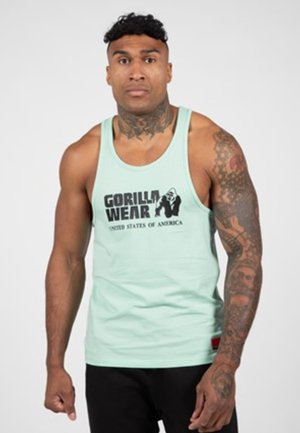 Gespierde man met tatoeages op de nek en rechterarm, gekleed in een lichtblauw, mouwloos Gorilla Wear tanktop en zwarte broek.