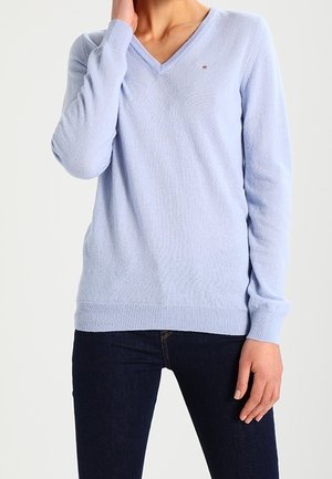 Femme portant un pull col en V bleu clair et un jean bleu foncé, main touchant le côté du cou, debout devant un fond uni.