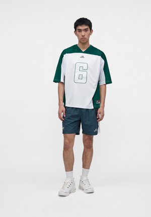 Junger Mann trägt ein grün-weißes Sporttrikot mit der Nummer 6, dunkle Shorts, weiße Socken und weiße Sportschuhe, steht vor einfachem Hintergrund.