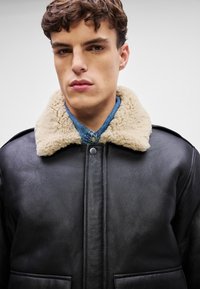 Black leather jacket with a beige shearling collar, featuring large front pockets, a zip closure, and a denim shirt underneath.