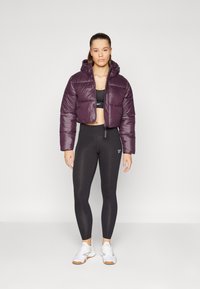 Giacca a vento cropped viola con cappuccio, abbinata a un reggiseno sportivo nero e leggings a vita alta. Le scarpe da ginnastica bianche completano il look.