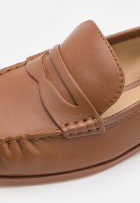 Mocassins en cuir marron avec un bout rond, des détails cousus et une texture lisse avec un discret accent en relief sur la partie supérieure.