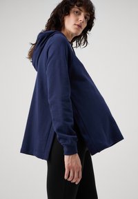 Sudadera con capucha de color navy, de corte relajado, con mangas largas, aberturas laterales y una textura suave. Combinada con leggings negros.