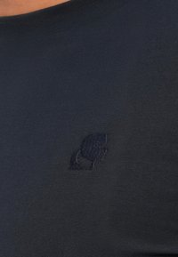 Mørkeblått stoff med en liten brodert silhuett av en persons profil og bundet hår i matchende tråd farge.