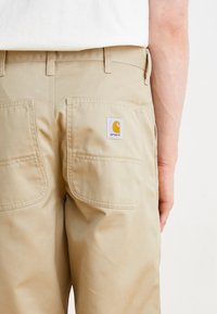 Beige work pants made of durable fabric with a straight cut, featuring two back pockets and a square logo patch in orange and white.