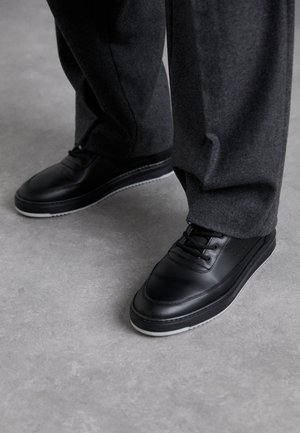 Person wearing black leather sneakers with white soles and dark grey pants standing on a smooth grey floor.