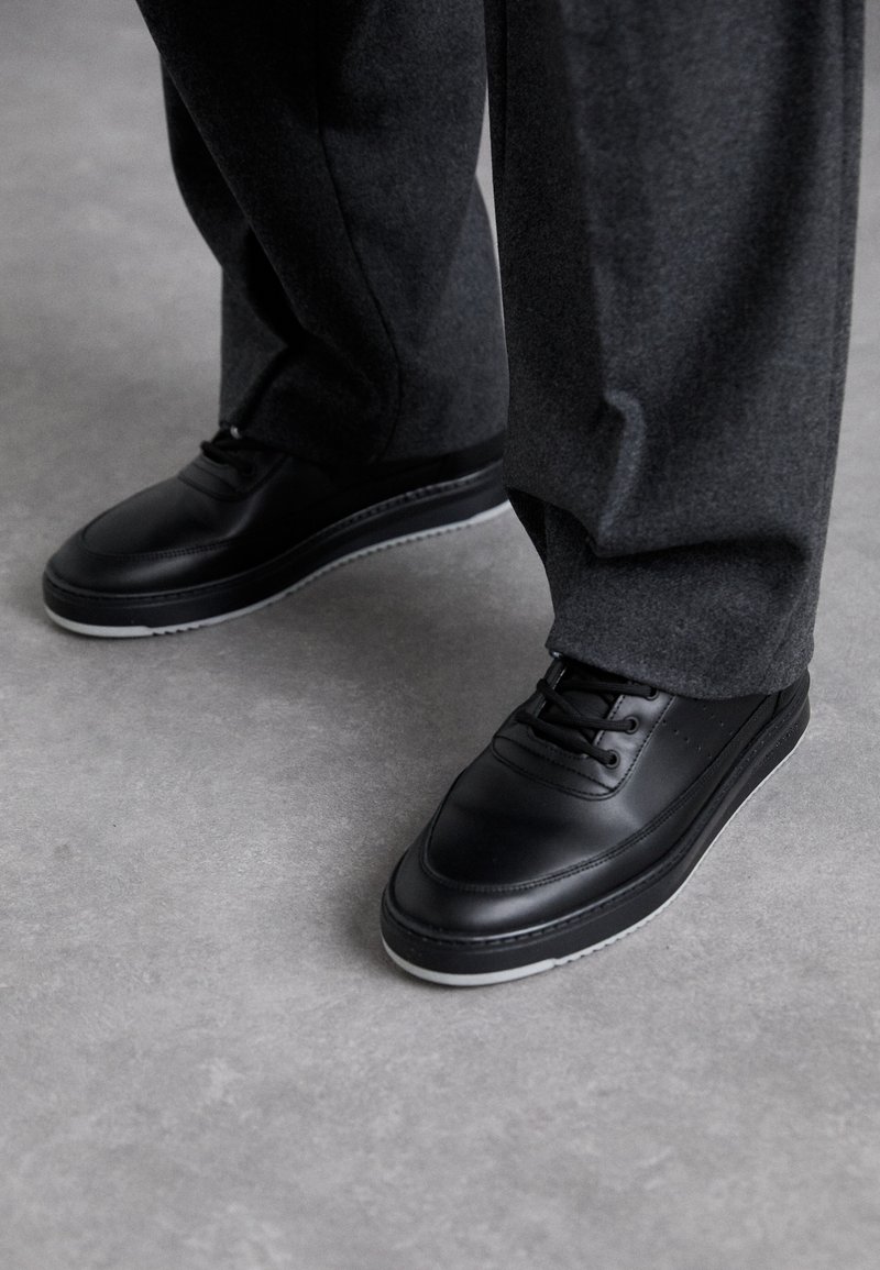 Person med svarta lädersneakers med vita sulor och mörkgrå byxor som står på en jämn grå golv.