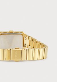 Goldfarbene Fossil-Uhr mit rechteckigem Zifferblatt, metallischem Armband mit gebürsteten und polierten Gliedern sowie einem Verschluss, der mit dem Markennamen versehen ist.