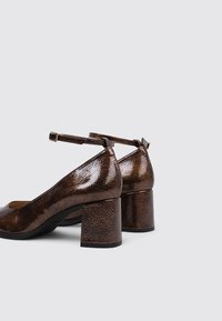 Zapatos de charol marrón con tacón cuadrado, punta redonda y correa al tobillo. Superficie texturizada con acabado brillante y diseño de tacón resistente.