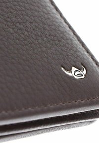 Gros plan sur le coin d'un portefeuille en cuir brun texturé avec un petit emblème argenté représentant un "G" stylisé et des formes courbes.