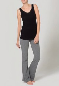 Kvinne som står barbeint iført en svart ermeløs topp med blonder og grå flagrende bukser mot en ensfarget lys bakgrunn.