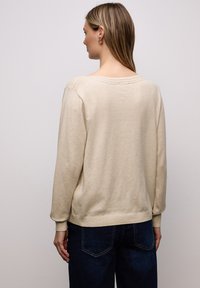 Pull beige en tricot avec encolure ronde, accents côtelés aux épaules et aux poignets, et coupe décontractée. Associé à un jean bleu foncé.