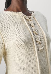 Pull en tricot couleur crème avec une surface texturée et un devant à lacets doté de quincaillerie de couleur argentée et d'un col décontracté.
