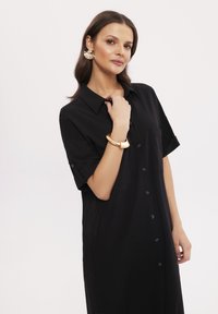 Czarna sukienka koszulowa z krótkimi rękawami, kieszonką na piersi i luźnym krojem, wykonana z gładkiego materiału o minimalistycznym designie.