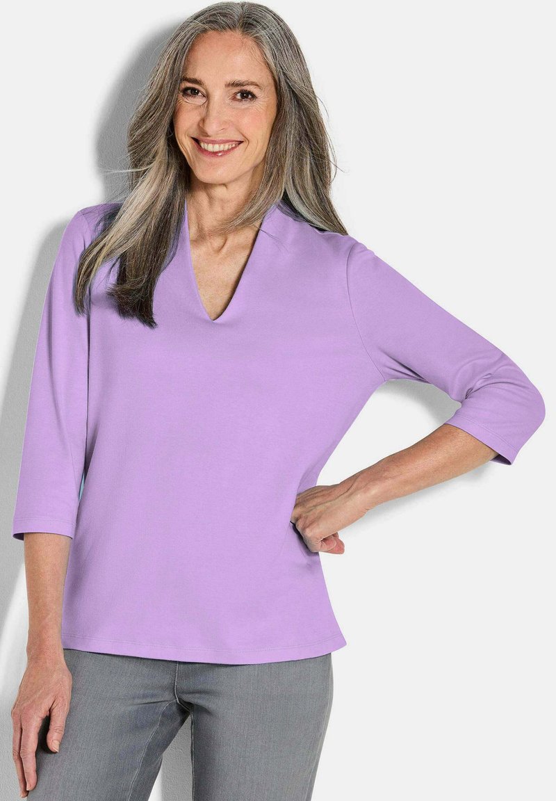 Lilafarbener V-Ausschnitt-Oberteil mit 3/4-Ärmeln, das eine glatte Textur und einen leicht ausgestellten Saum aufweist. Kombiniert mit hellgrauen Hosen.