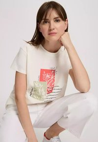 Kremowa koszulka z krótkim rękawem, z botanicznymi nadrukami w czerwieni i zieleni oraz tekstem w czerni. Białe, krótkie spodnie i buty z czerwonym akcentem.