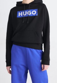 Sudadera con capucha negra con un logo rectangular azul de "HUGO", con un bolsillo frontal, combinada con pantalones de chándal azules. Tela suave, ajuste relajado.